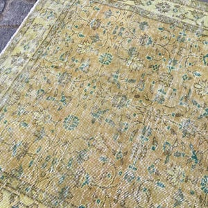 May include: An antique, distressed rug with a faded, floral pattern. The rug features a warm, golden-yellow base color with accents of teal and brown. The design includes stylized flowers and vines, creating a vintage aesthetic.