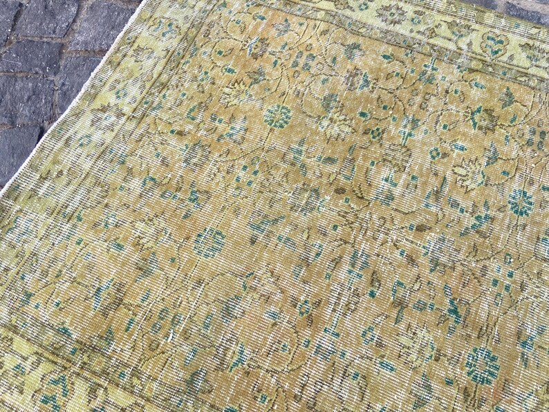 May include: An antique, distressed rug with a faded, floral pattern. The rug features a warm, golden-yellow base color with accents of teal and brown. The design includes stylized flowers and vines, creating a vintage aesthetic.