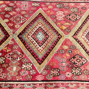 May include: A woven rug featuring a vibrant red background with large, repeating diamond-shaped patterns. The diamonds contain intricate geometric motifs in shades of yellow, teal, black, and white, interspersed with smaller floral-like shapes.