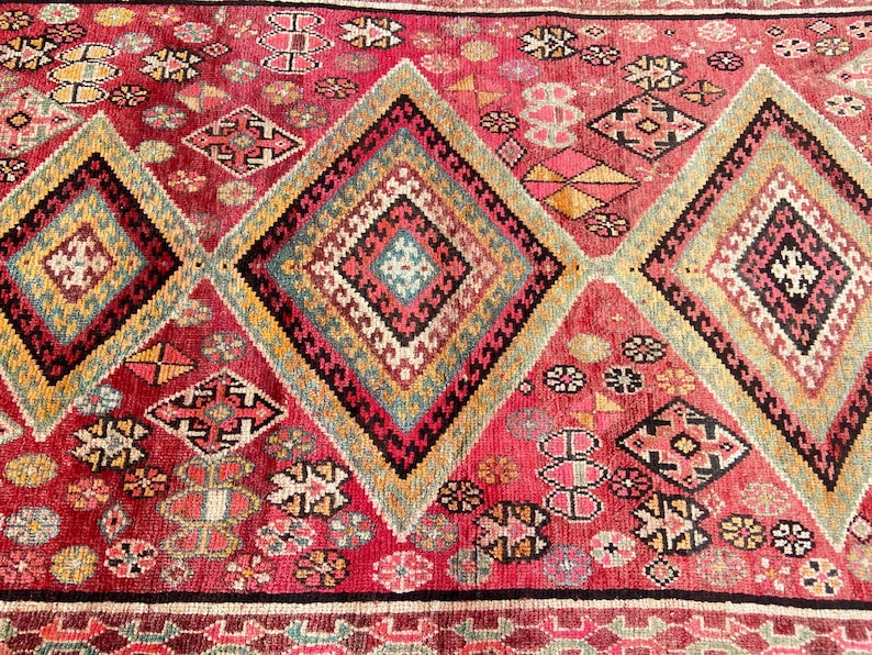 May include: A woven rug featuring a vibrant red background with large, repeating diamond-shaped patterns. The diamonds contain intricate geometric motifs in shades of yellow, teal, black, and white, interspersed with smaller floral-like shapes.