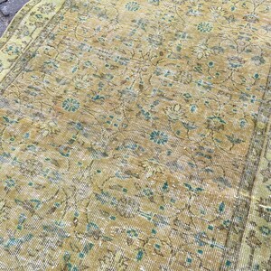 May include: Close-up of a distressed, vintage rug with a faded, floral pattern. The rug features a color palette of muted gold, teal, and brown. The design includes stylized floral motifs and a border.