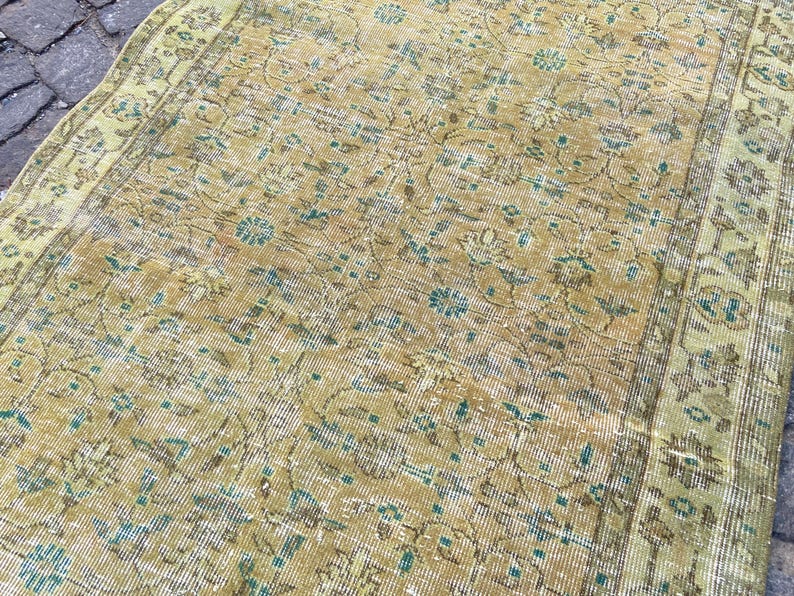 May include: Close-up of a distressed, vintage rug with a faded, floral pattern. The rug features a color palette of muted gold, teal, and brown. The design includes stylized floral motifs and a border.