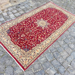 May include: A red rug with a floral pattern and a cream-colored border. The rug has a traditional design and is made of wool.