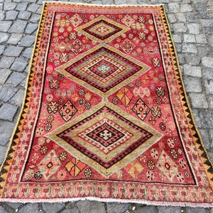 May include: A rectangular area rug featuring a vibrant red background with three large central diamond motifs in shades of olive green, yellow, and black. The rug is decorated with intricate geometric patterns and a serrated border in black and yellow.