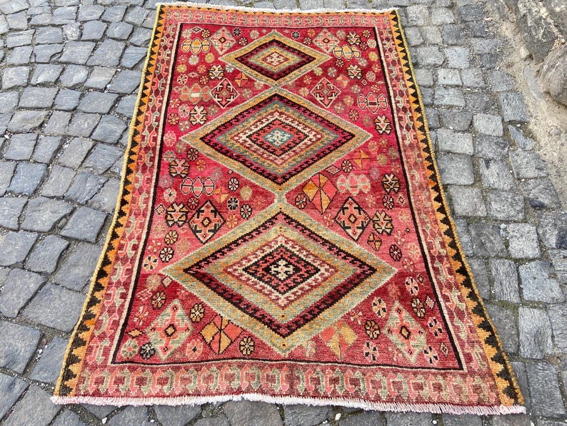 May include: A rectangular area rug featuring a vibrant red background with three large central diamond motifs in shades of olive green, yellow, and black. The rug is decorated with intricate geometric patterns and a serrated border in black and yellow.