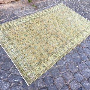 May include: A rectangular, vintage-style rug with a faded, floral pattern in shades of yellow, green, and beige. The rug has a detailed border and is placed on a cobblestone surface. The rug's dimensions are not visible.