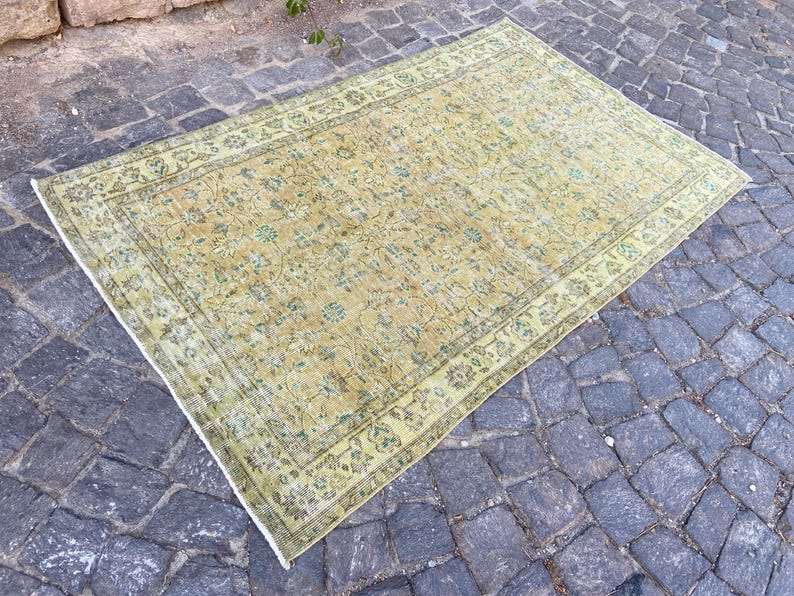 May include: A rectangular, vintage-style rug with a faded, floral pattern in shades of yellow, green, and beige. The rug has a detailed border and is placed on a cobblestone surface. The rug's dimensions are not visible.