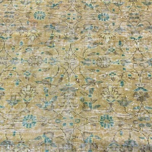 May include: Close-up of a vintage rug with a distressed appearance. The rug features a repeating floral pattern in shades of beige, brown, and teal. The design has a faded, antique look, with visible wear and tear.