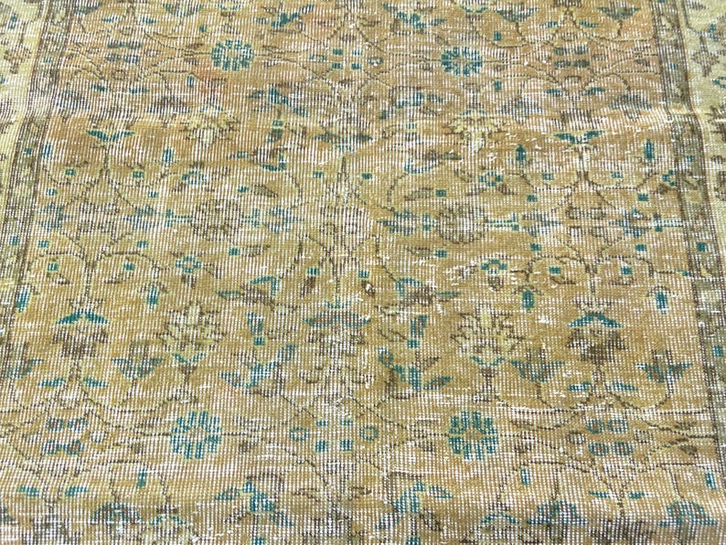 May include: Close-up of a vintage rug with a distressed appearance. The rug features a repeating floral pattern in shades of beige, brown, and teal. The design has a faded, antique look, with visible wear and tear.