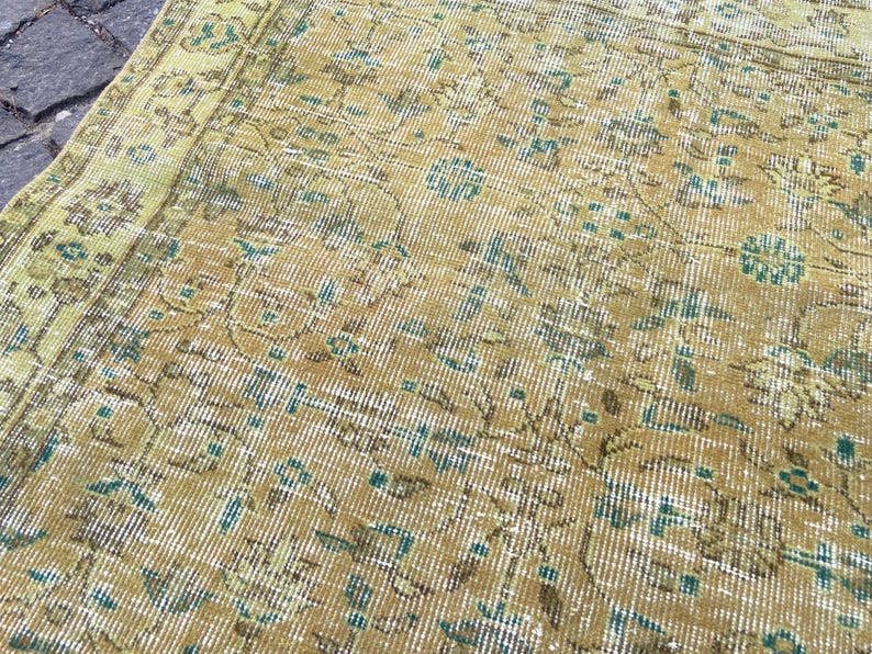 May include: Close-up of a vintage rug with a distressed, faded appearance. The rug features a warm color palette of gold, brown, and teal, with intricate floral and geometric patterns. The rug's design suggests a traditional, oriental style.