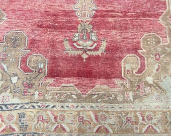 May include: Close-up of a vintage rug with a distressed, faded appearance. The rug features a central field of deep red, surrounded by intricate beige and brown geometric patterns. The border has a repeating design with muted colors.