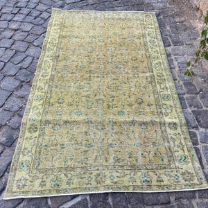 May include: A rectangular, vintage-style rug with a faded, distressed appearance. The rug features a central field of muted yellow and green tones with floral patterns. A border in a darker shade of green frames the design. The rug is placed on a cobblestone surface.