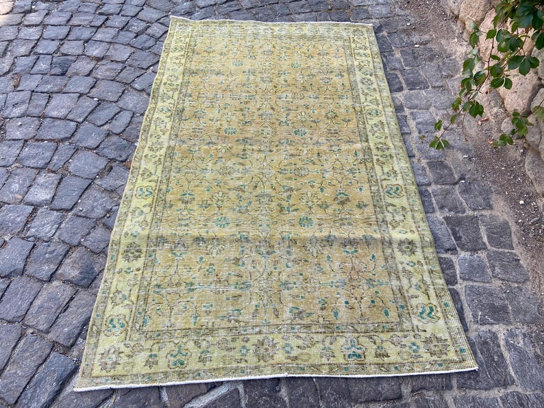 May include: A rectangular, vintage-style rug with a faded, distressed appearance. The rug features a central field of muted yellow and green tones with floral patterns. A border in a darker shade of green frames the design. The rug is placed on a cobblestone surface.