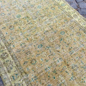May include: Vintage-style rug with a distressed appearance. The rug features a warm, golden-yellow base color with faded floral patterns in shades of teal and brown. The edges are bordered with a lighter green hue, creating a classic, aged aesthetic.