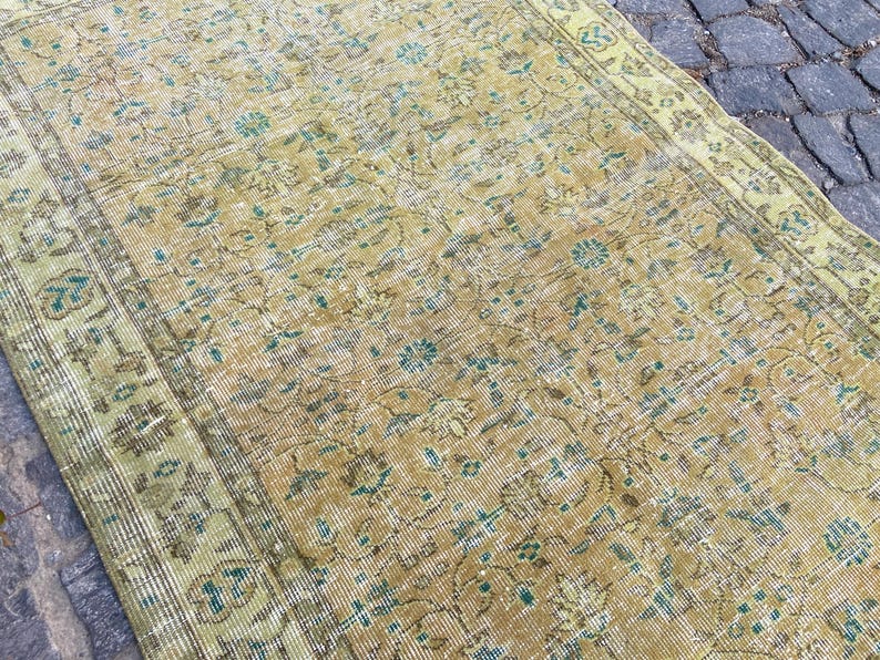 May include: Vintage-style rug with a distressed appearance. The rug features a warm, golden-yellow base color with faded floral patterns in shades of teal and brown. The edges are bordered with a lighter green hue, creating a classic, aged aesthetic.