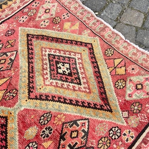 May include: A rectangular woven rug featuring a vibrant red base with large central diamond motifs in shades of yellow, black, and white. The surface is decorated with intricate geometric patterns and small floral-like shapes throughout the field.