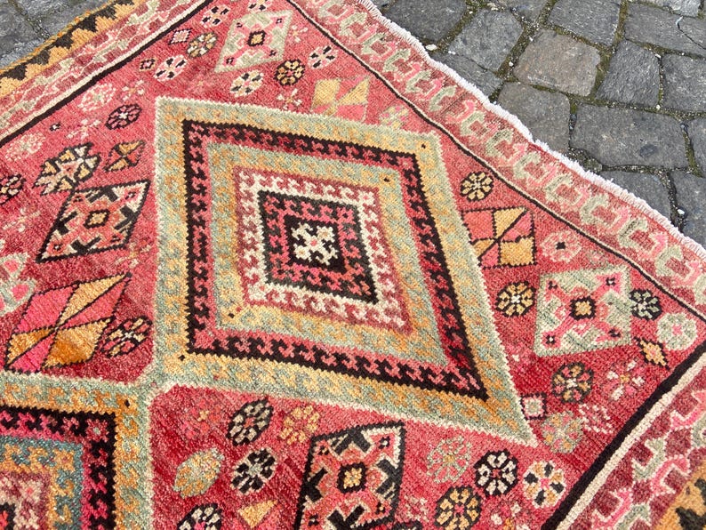 May include: A rectangular woven rug featuring a vibrant red base with large central diamond motifs in shades of yellow, black, and white. The surface is decorated with intricate geometric patterns and small floral-like shapes throughout the field.