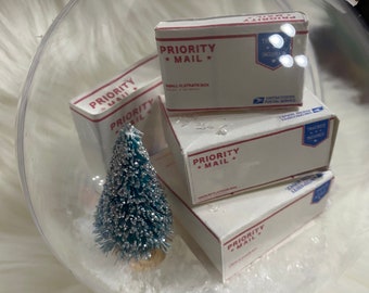 May include: A clear glass ornament with a miniature Christmas tree and four miniature Priority Mail boxes inside. The boxes are stacked on top of each other and have the words "Priority Mail" printed on them.