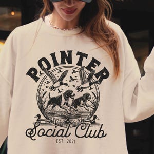 May include: A beige sweatshirt with a black graphic print of a hunting dog and the text "POINTER SOCIAL CLUB EST. 2021".