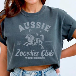 May include: A dark grey cropped t-shirt with a white graphic of an Australian Shepherd running. The graphic includes the text "Aussie" and "Zoomies Club" with the tagline "Watch your six!"