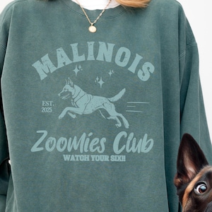 May include: Teal sweatshirt featuring the text "Malinois Zooomies Club" and a graphic of a running dog. The sweatshirt also has the text "Est. 2025" and "Watch Your Six!!". A happy Malinois dog is on the right.