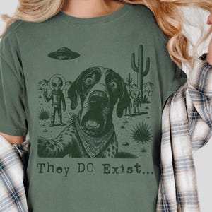 May include: Green t-shirt with a graphic of a dog taking a selfie with aliens and a UFO. The text "They DO Exist..." is printed below the image. The dog is wearing a bandana, and the background features desert scenery.
