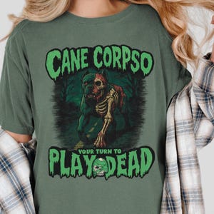 May include: Green t-shirt with the text "CANE CORPSO" in a green, spooky font. Below the text is a graphic of a dog skeleton. The text "YOUR TURN TO PLAY DEAD" is below the graphic in a green font.