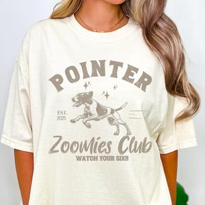 May include: A cream-coloured t-shirt with a graphic design. The design features the word "POINTER" above a running dog illustration. Below the dog, the text reads "Zoomies Club" and "Watch Your Six!!". The shirt also has "EST. 2025".