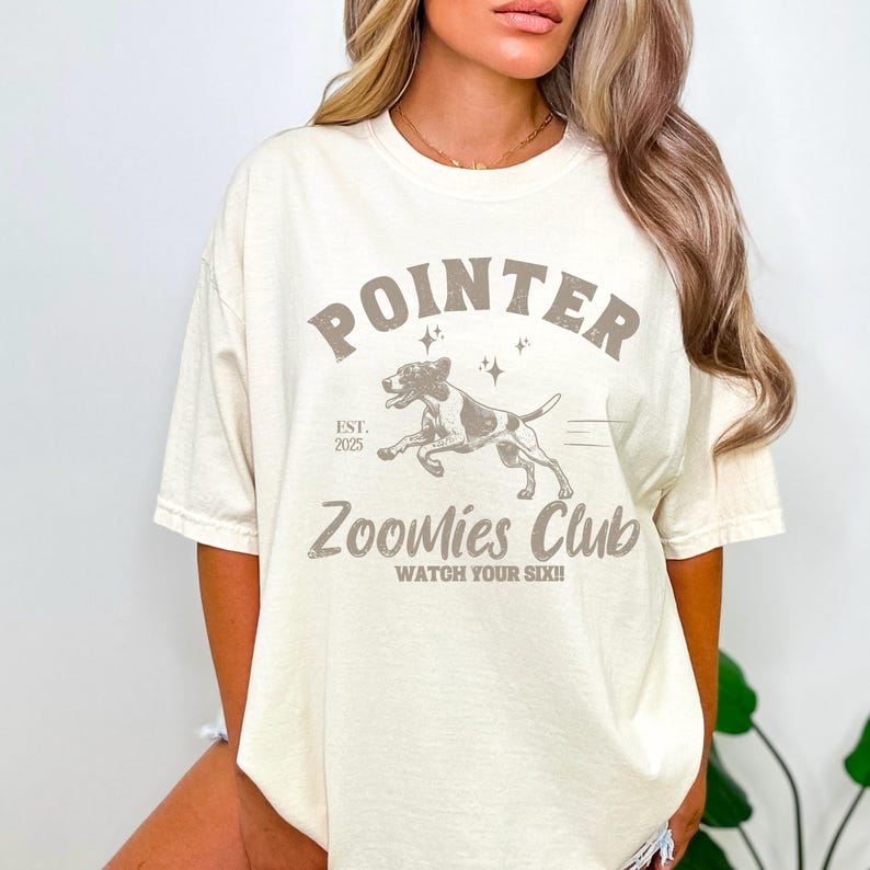 May include: Cream-colored t-shirt with a graphic design featuring the word "POINTER" above a running dog illustration. Below the dog, the text reads "Zoomies Club" and "Watch Your Six!!". The shirt also has "EST. 2025".