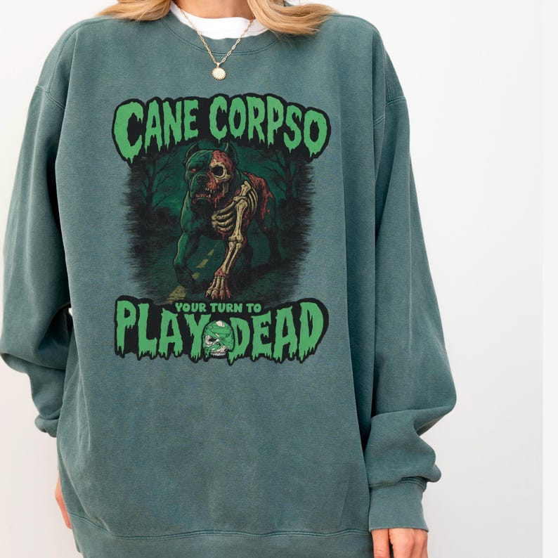 May include: Teal green sweatshirt with the text "CANE CORPSO" above a graphic of a zombie dog. Below the dog is the text "PLAY DEAD" with a skull graphic. The sweatshirt is a crew neck style.