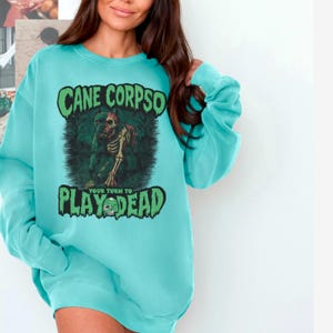 May include: A turquoise sweatshirt with the text "CANE CORPSO" above a graphic of a skeleton dog and the words "PLAY DEAD". The sweatshirt is a solid color with long sleeves.