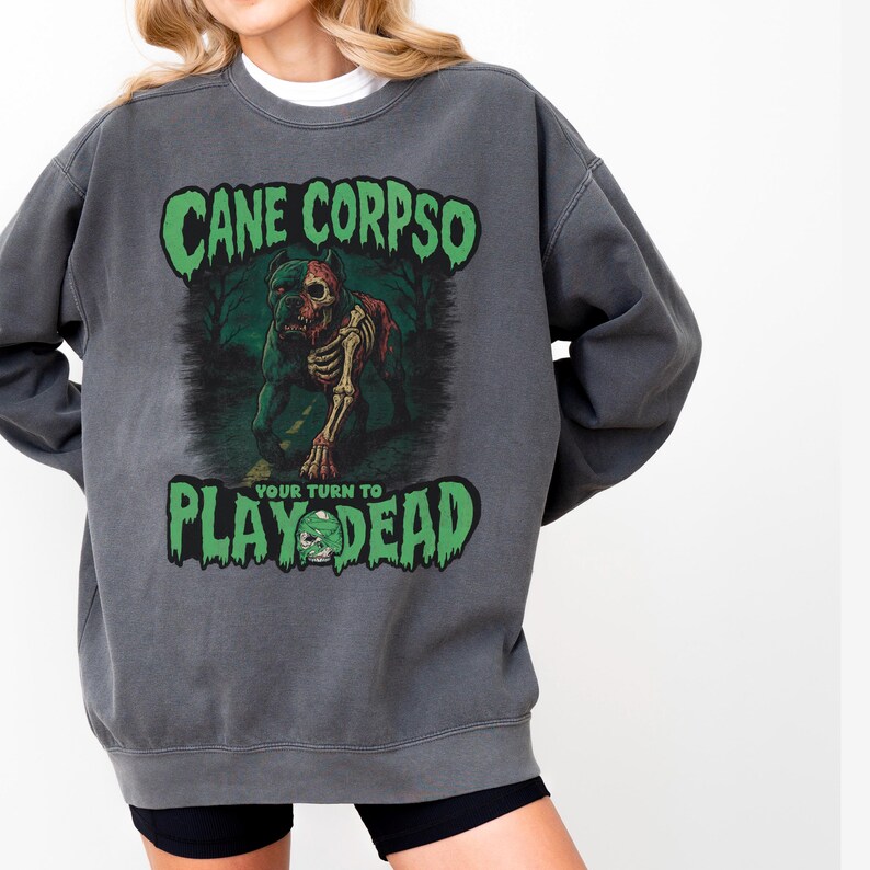 May include: Dark gray sweatshirt with the text "CANE CORPSO" above a graphic of a zombie dog and the text "PLAY DEAD". The graphic is in green, red, and white. The sweatshirt has a crew neck.