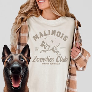 May include: Cream t-shirt with the text "Malinois Zoomies Club" and a running dog graphic. The shirt also features the text "Watch Your Six!!" and "Est. 2025". The shirt is worn with a brown plaid shirt.