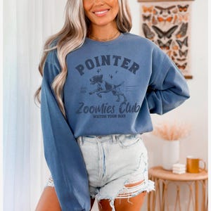 May include: A blue long-sleeve sweatshirt with the text "POINTER" above a graphic of a dog and the words "Zoomies Club". The sweatshirt is a solid blue color and has a vintage style.