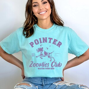 May include: A light blue t-shirt with a purple graphic that reads "POINTER" above a running dog illustration. Below the dog is "Zoomies Club" and "Watch Your Six!!". The shirt also has "Est. 2025" printed on it.