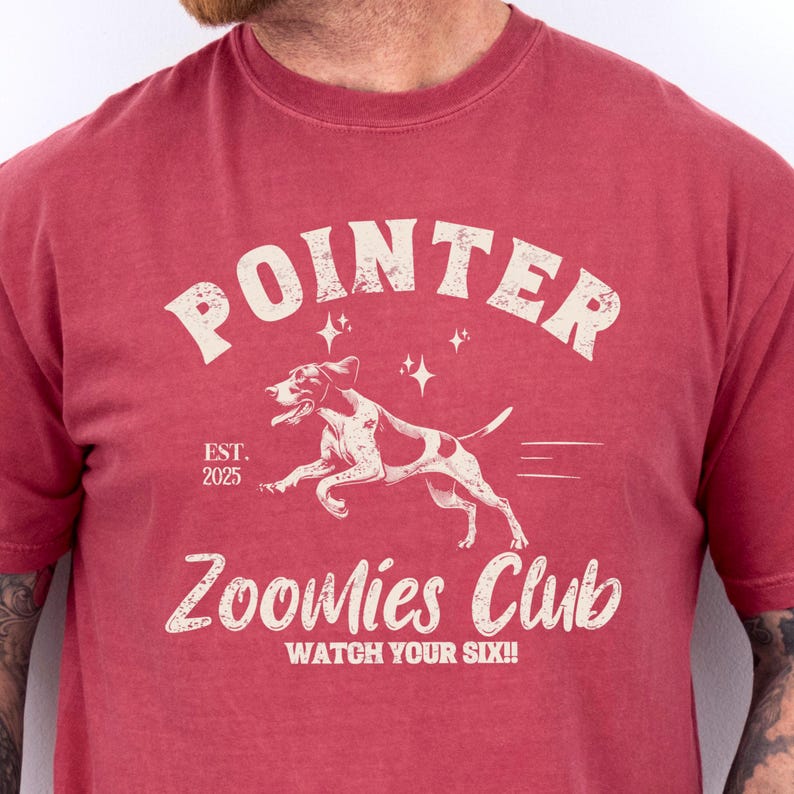 May include: Red t-shirt with a graphic featuring a running dog, the word "POINTER" above, and "Zoomies Club" below. The text "EST. 2025" and "WATCH YOUR SIX!!" are also included. The dog is white with brown spots.