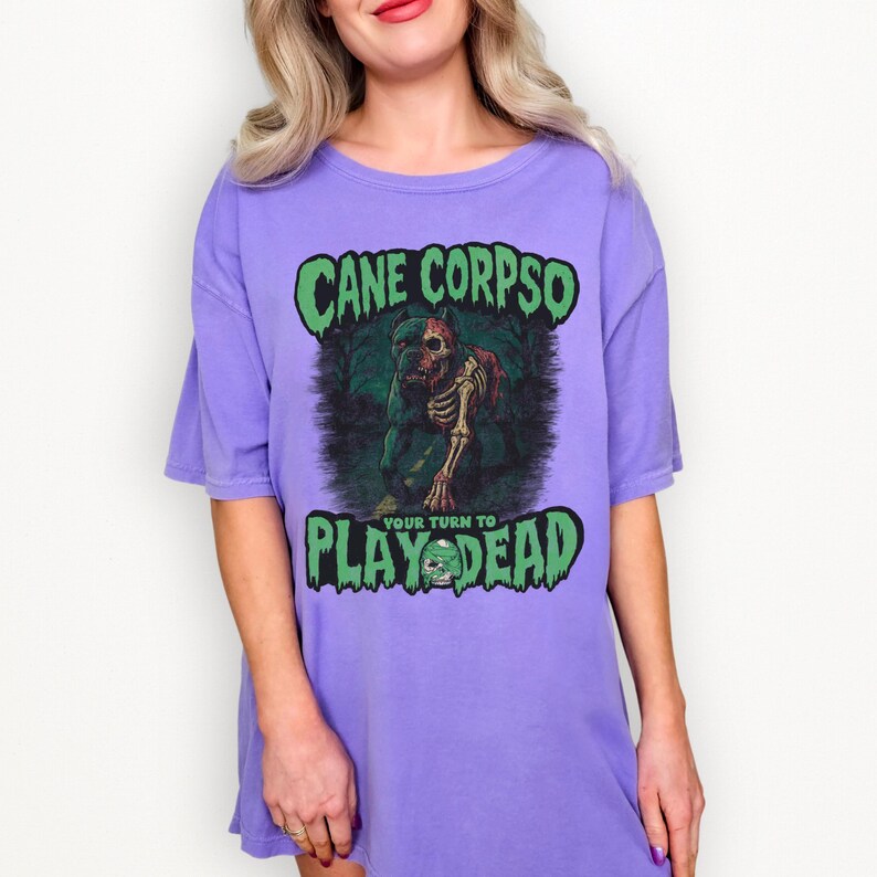 May include: A lilac-colored t-shirt dress with a graphic featuring a skeletal dog and the text "CANE CORPSO" and "PLAY DEAD". The design is in green and brown tones.