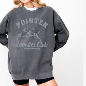 May include: Dark gray sweatshirt with the word "POINTER" above a graphic of a dog and the text "Zoomies Club". The sweatshirt has a crew neck and long sleeves. The text "Watch Your Six!!" is below the dog graphic.