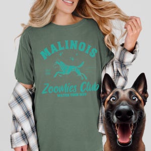 May include: Green t-shirt featuring the text "Malinois Zoomies Club" and a teal running dog graphic. The shirt also has the text "Watch Your Six!". A smiling Belgian Malinois dog is in the image.