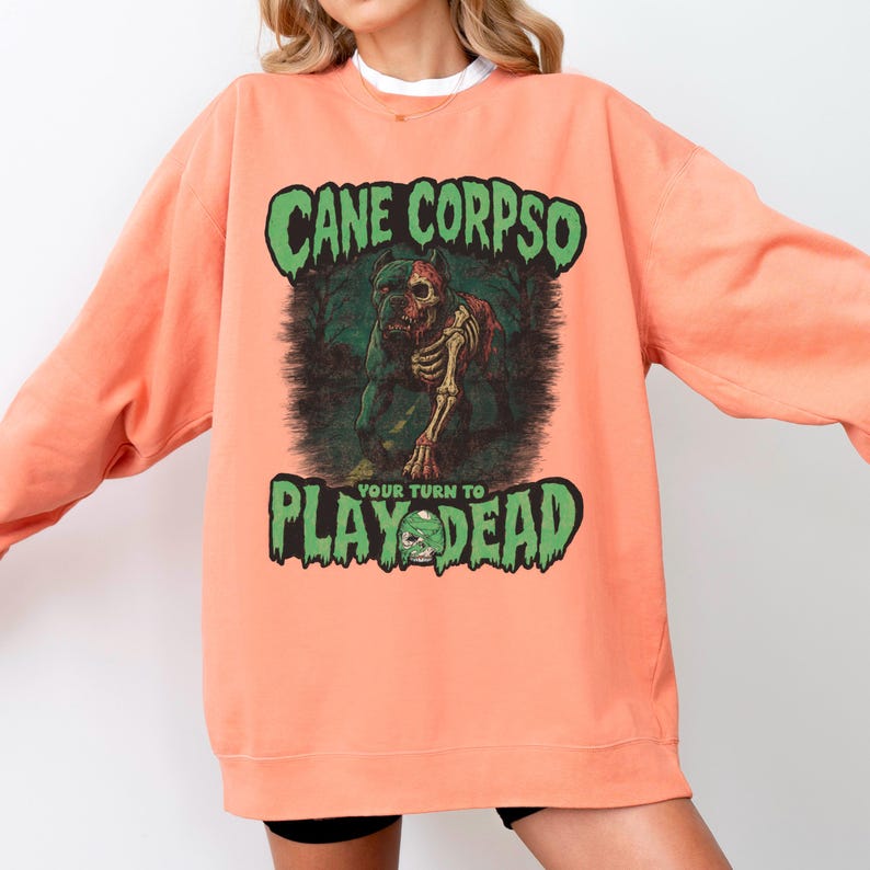 May include: Peach-colored sweatshirt with a graphic featuring a zombie dog and the text "Cane Corposo, Your Turn to Play Dead" in green. The design includes a forest background and a skull.