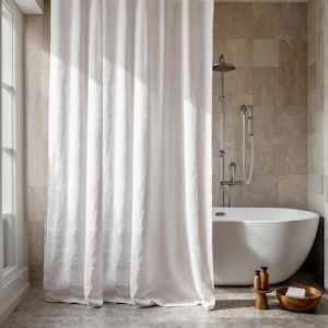 May include: A white linen shower curtain hangs in front of a white bathtub. The bathroom has a neutral color palette with stone-like tiles and a wooden side table with bath products.