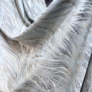 Könnte beinhalten: Ein Stoff mit einem fließenden, abstrakten Muster in Hellblau, Silber und Weiß. Das Design besteht aus wellenförmigen Linien, die eine Bewegung über die Oberfläche erzeugen, ergänzt durch einen dezenten metallischen Glanz im gesamten Gewebe.
