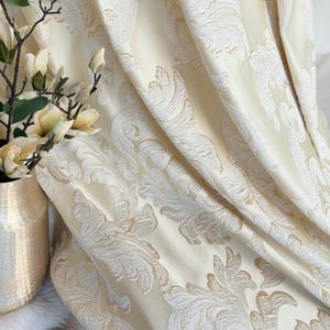 May include: Cream-colored damask curtain fabric with a large-scale, scrolling floral pattern in a lighter cream color. The fabric is draped, showing the texture and design. A gold vase with flowers is in the background.