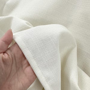 K&ouml;nnte beinhalten: Nahaufnahme eines cremefarbenen Stoffes mit strukturiertem Gewebe. Der Stoff wird gehalten, wodurch sein Fall und die subtilen Variationen seiner Oberfl&auml;che sichtbar werden. Das Material wirkt weich und hat einen nat&uuml;rlichen, cremefarbenen Farbton.