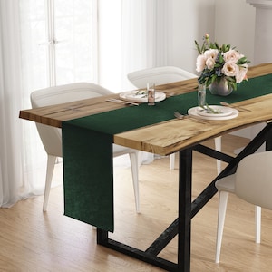 May include: A dark green table runner on a wooden dining table. The table has a live edge design and black metal legs. White chairs are positioned around the table, and a vase of flowers sits in the centre. The table is set with plates and cutlery.