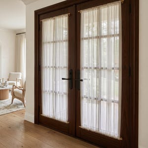 May include: A pair of dark brown wooden French doors featuring glass panes covered by sheer, vertical-striped white curtains. The curtains are held in place by thin gold-toned rods at the top and bottom of each glass panel, with black handles.