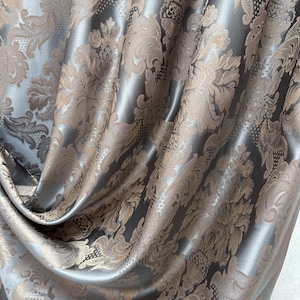 May include: A fabric curtain featuring a repeating damask pattern in shades of metallic silver and muted bronze. The material has a smooth, reflective sheen with deep folds that create a sense of volume and texture across the surface.