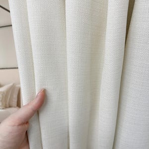 K&ouml;nnte beinhalten: Nahaufnahme von cremefarbenen Leinen-Vorh&auml;ngen mit strukturiertem Gewebe. Der Stoff drapiert sich in weichen Falten und zeigt die subtile Textur und Farbvariationen. Die Vorh&auml;nge scheinen aus einem strapazierf&auml;higen, hochwertigen Material zu bestehen, das f&uuml;r die Inneneinrichtung geeignet ist.