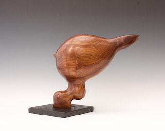 May include: A wooden sculpture of a bird-like form, with a smooth, polished finish. The sculpture is a warm brown color and is mounted on a dark square base. The bird's head is tilted upwards, and its body curves gracefully.
