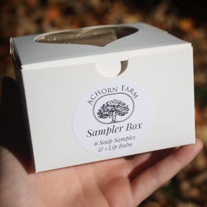May include: A white sampler box with a heart-shaped cutout on top, held in a hand. The box features a circular label with a tree illustration and the text "ACHORN FARM Sampler Box 6 Soap Samples & 1 Lip Balm."